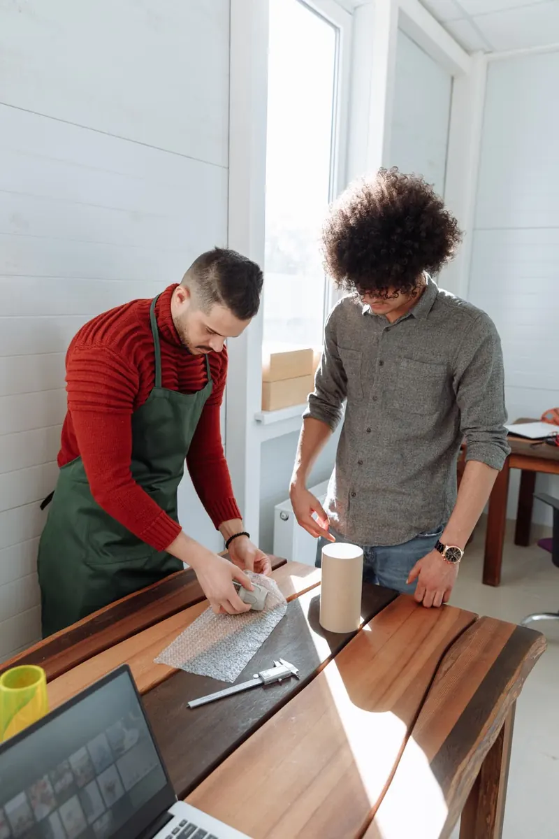Dos personas preparan paquetes en una mesa de trabajo junto a un portátil