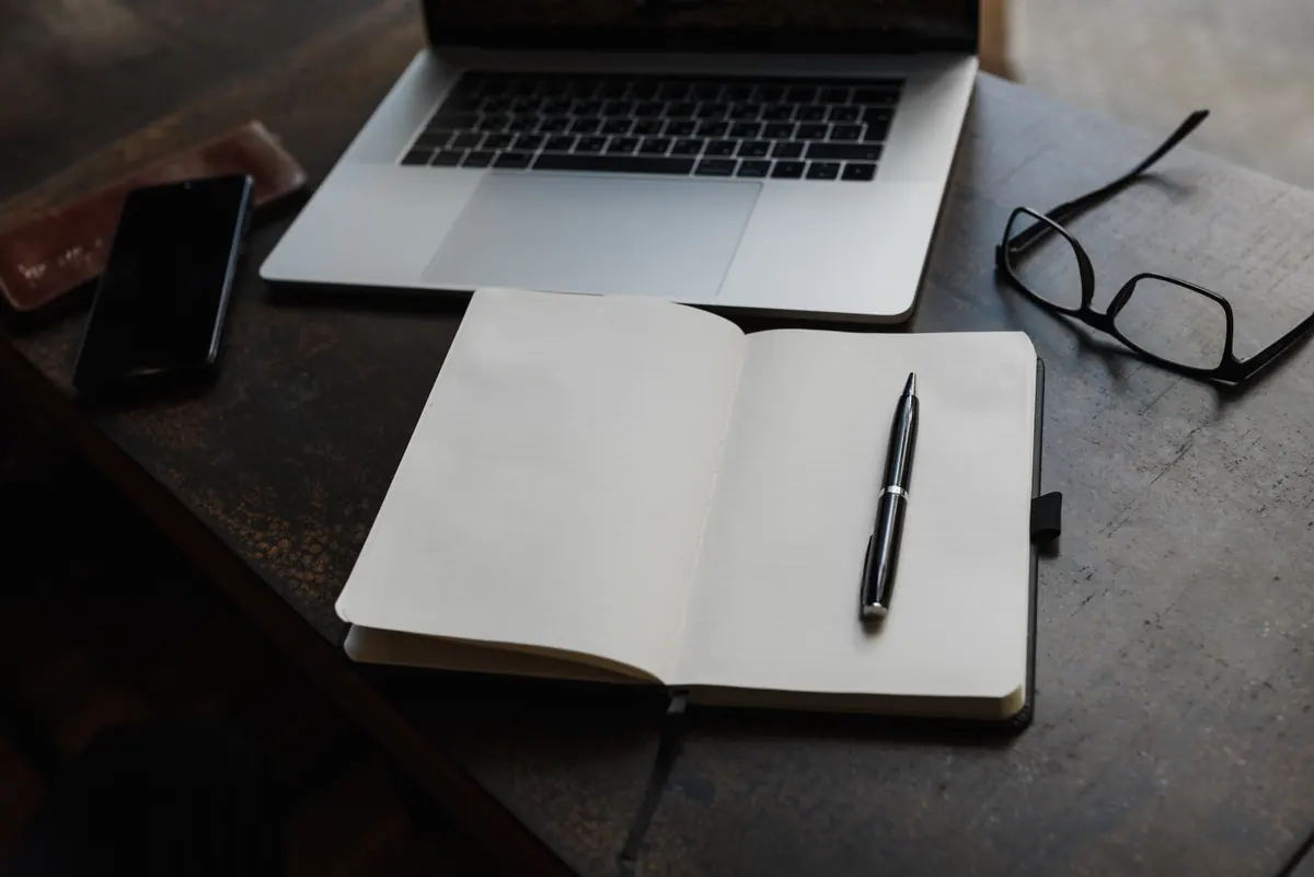 Cuaderno abierto con bolígrafo junto a un portátil y unas gafas