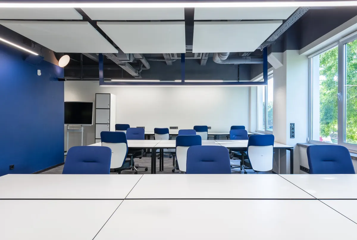 Sala de trabajo vacía con mesas blancas y sillas azules junto a ventanas